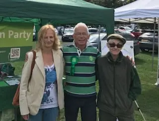 Three people standing in front of the Green Party stall at an outdoor market.