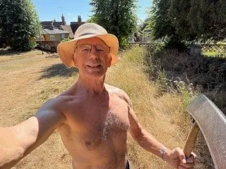 Edward Gildea in a sun hat, holding his scythe in a churchyard of long dry grass on a sunny day