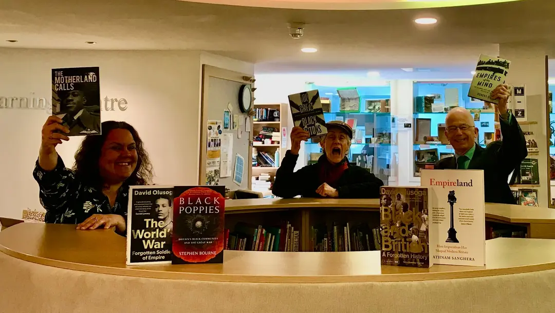 Three people behind a library counter holding books in the air, with more books stood along the counter.