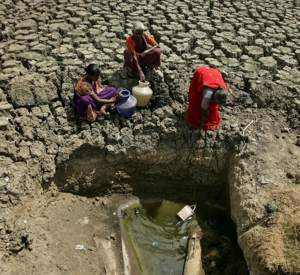 Women collecting water in pots from a small pool at the bottom of a dried-up, cracked riverbed in India