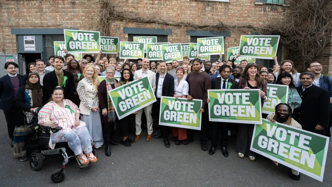 A large group of Green Party candidates and campaigners outside a brick building, holding up “Vote Green” placards