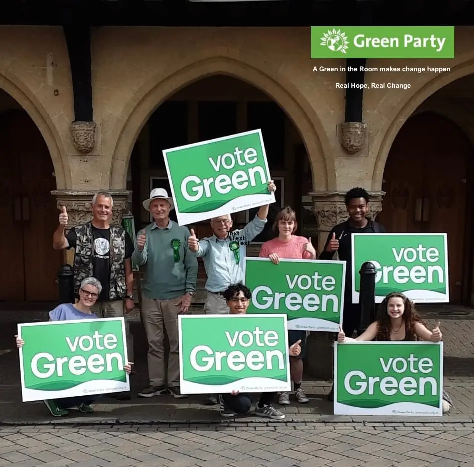 Nine people in front of stone arches holding “Vote Green” placards, several giving a thumbs up. A Green Party logo and the words “A Green in the Room makes change happen. Real Hope, Real Change” are printed in one corner.