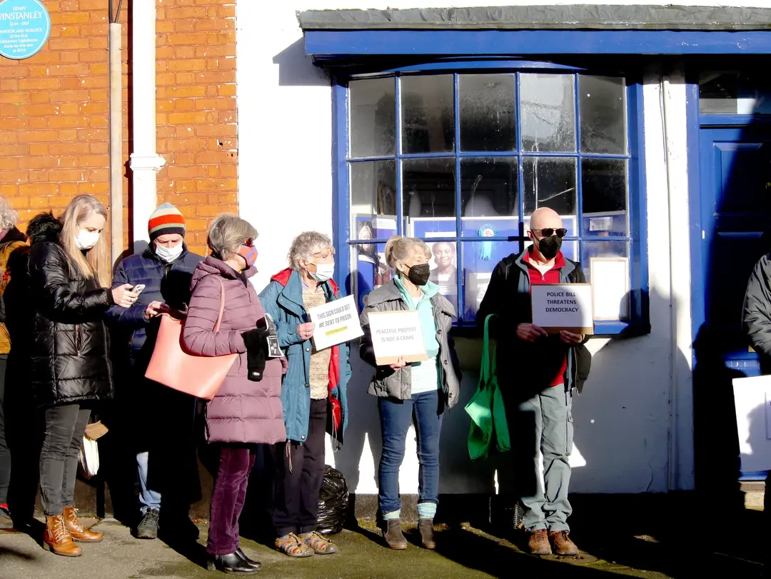 People in winter coats and face masks holding placards beside a blue shop window. One placard reads “Peaceful protest is not a crime”.
