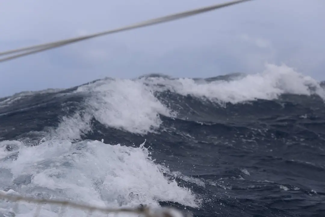 A large wave breaking at sea under a grey sky.